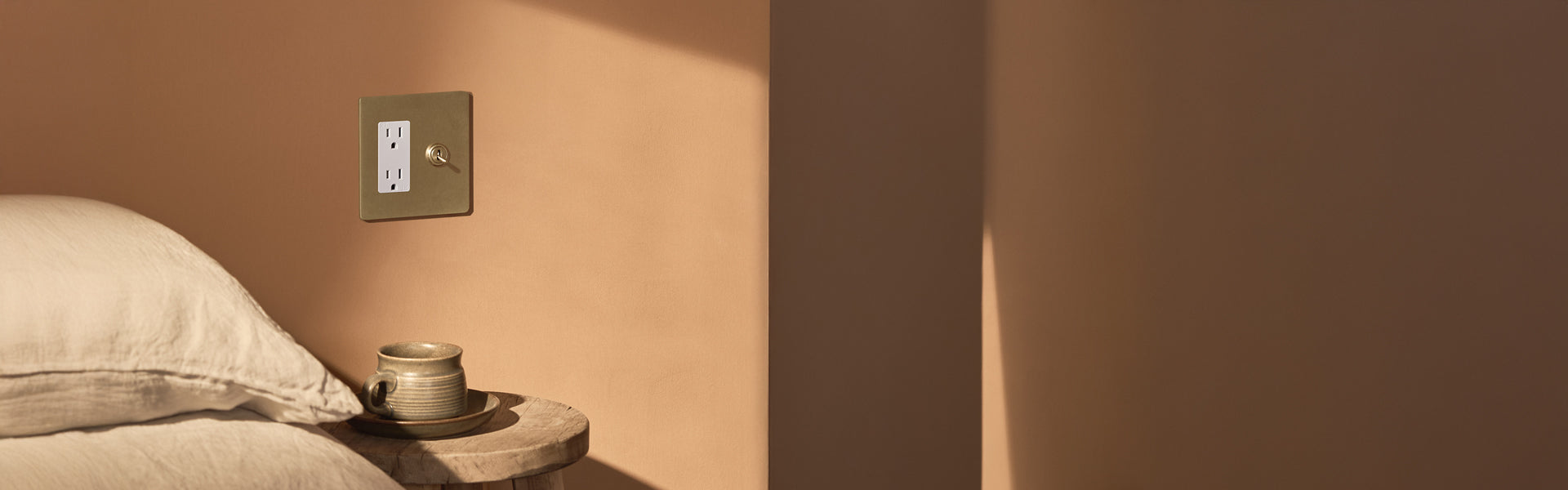 A minimalist bedside scene featuring a clay cup on a wooden table, with a wall switch above a peach-colored wall. Soft bedding is visible.