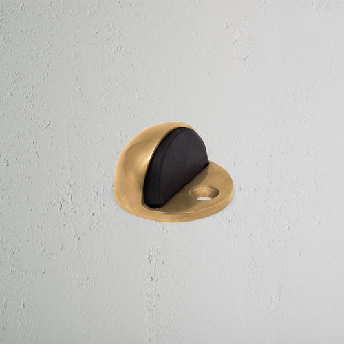 Clifton Half Moon Door Stop in antique brass with black rubber insert on a light gray textured wall.