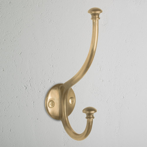 Mews Hook in antique brass, a wall-mounted coat hook with two curved arms on a light gray wall.
