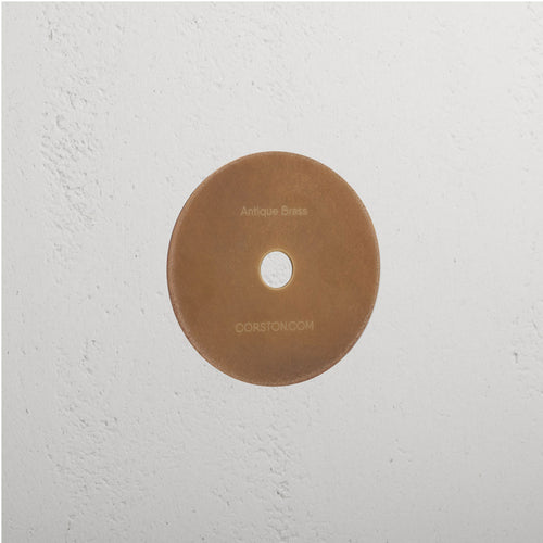 Circular Sample Finish in Antique Brass with central hole, mounted on white textured wall, labeled 'CORSTON.COM'.