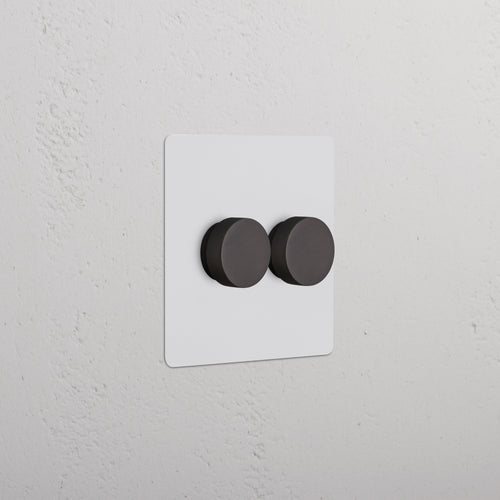 Kew 2G Dimmer Switch in paintable bronze with two round black knobs, shown on a wall.