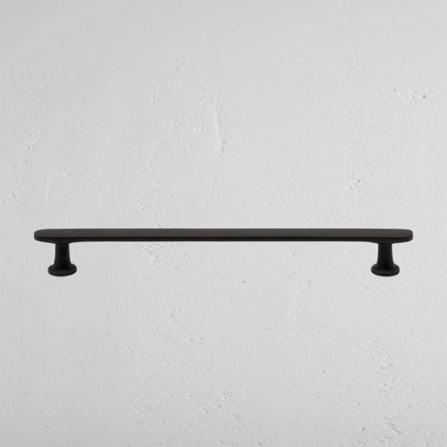Milliner Furniture Handle Medium in bronze with rounded ends, mounted on a white textured wall.