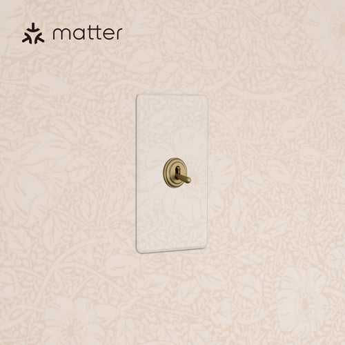 Kew 1G Smart Retractive Toggle Switch in Clear Antique Brass with clear faceplate on a cream floral wall.