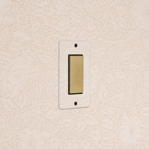 Kew 1G Center Retractive Rocker Switch in clear antique brass black on a wall with floral patterned wallpaper.