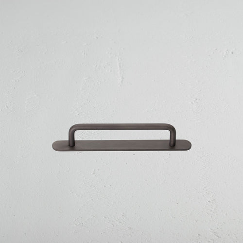 Small bronze Kilburn furniture handle with rounded ends, mounted horizontally on a textured white wall.