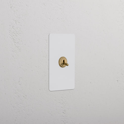 Kew 1G Toggle Switch in paintable antique brass with a white plate, mounted on a textured white wall.