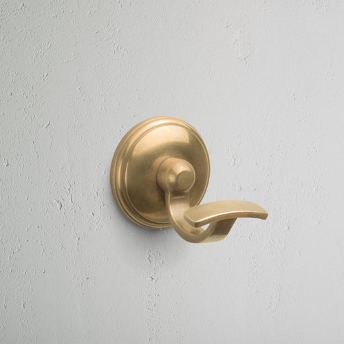 Antique brass Portland Hook with round base, mounted on a light gray textured wall.