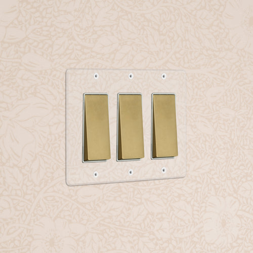 Kew 3G Rocker Switch in clear antique brass white, wall-mounted against beige floral-patterned wallpaper.
