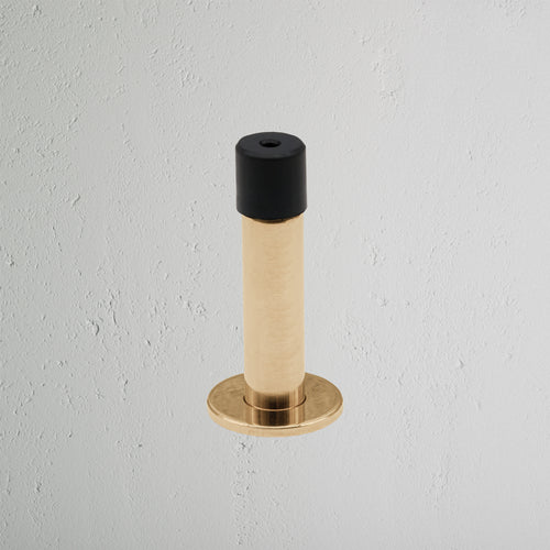 Montpelier Door Stop in antique brass finish with black rubber tip, mounted on a light gray wall.
