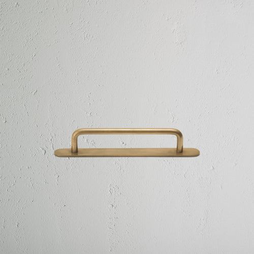 Kilburn Furniture Handle Small in antique brass, mounted horizontally on a textured white wall.