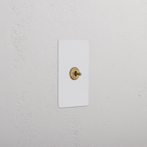 Kew 1G Center Retractive Toggle Switch in paintable antique brass with white plate, mounted on a white wall.