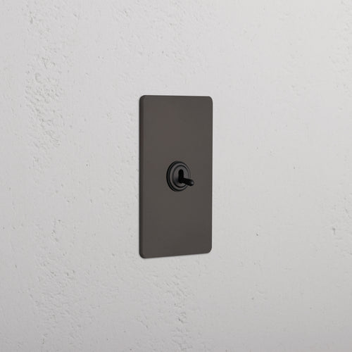 Bronze Grove 1G Retractive Toggle Switch with a rectangular plate installed on a white textured wall.