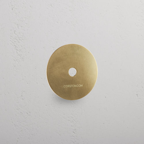 Circular Sample Finish in Antique Brass with central hole, mounted on white textured wall, labeled 'CORSTON.COM'.
