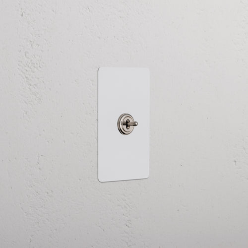Kew 1G Center Retractive Toggle Switch in paintable polished nickel, mounted on a white textured wall.