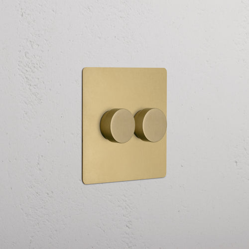 Kew 2G Dimmer Switch in Antique Brass with two round knobs, mounted on a white textured wall.