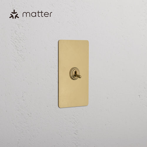 Kew 1G Smart Retractive Toggle Switch in Antique Brass, mounted on a white textured wall.