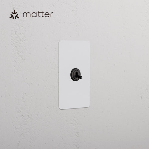 Kew 1G Smart Retractive Toggle Switch in paintable bronze finish with black toggle, shown on white wall.