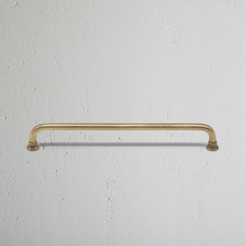 Medium Sycamore Furniture Handle in antique brass finish, shown mounted on a white textured surface.