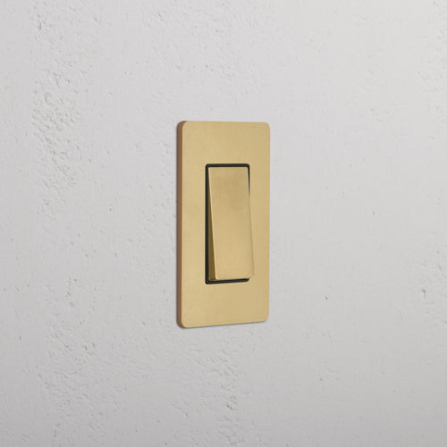 Grove 1G Rocker Switch in Antique Brass Black with a gold-tone rectangular plate on a white textured wall.