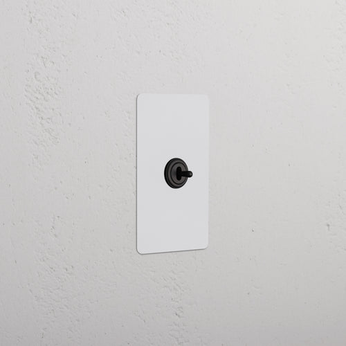 Kew 1G Center Retractive Toggle Switch in paintable bronze finish, single gang, on a white wall.