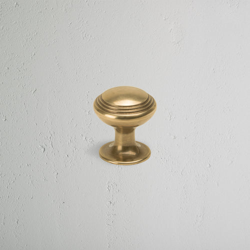 Small round Barlow furniture knob in antique brass with a smooth, stepped design on a light gray wall.