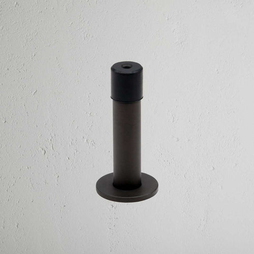 Montpelier Door Stop in bronze with a round base and black rubber tip mounted on a white wall.