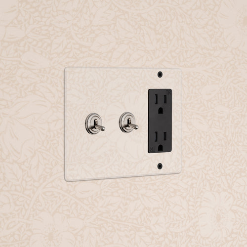 Kew 3G 2x Toggle Switch & Outlet in clear polished nickel black finish, mounted on floral wallpaper.